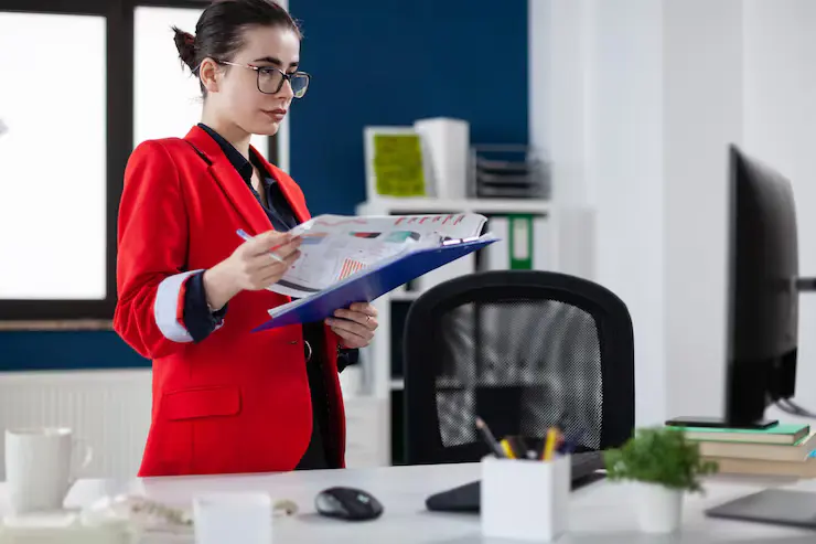 businesswoman-standing-up-desk-holding-clipboard-with-chart-focused-entrepreneur-with-glasses-red-jacket-startup-office-looking-business-results-computer-screen_482257-30235